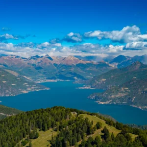 Aerial view of Lake Como with a forested ridge in the foreground, looking toward the peninsula of Bellagio nestled between two branches of the lake.