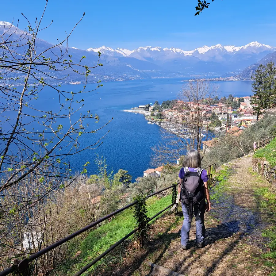 Cobblestone path winding down toward Bellano, with a female hiker enjoying the spectacular lake and snow-covered mountain views.