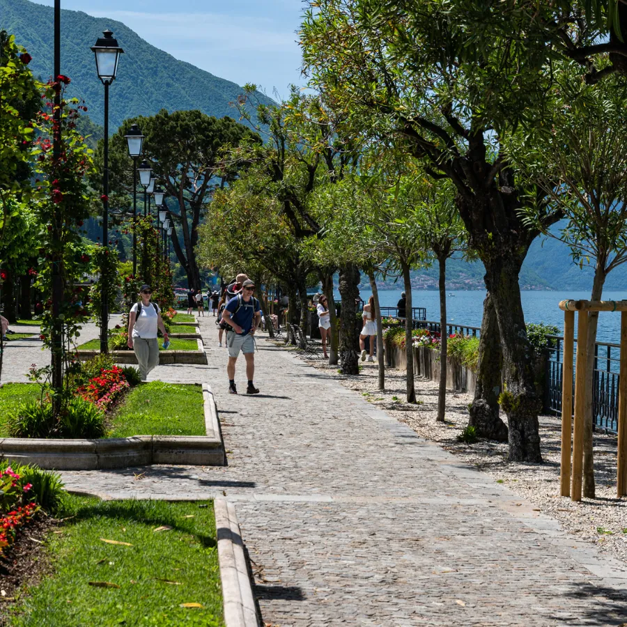 Tourists walking along the scenic lakeside promenade in Bellagio, lined with trees and flowers