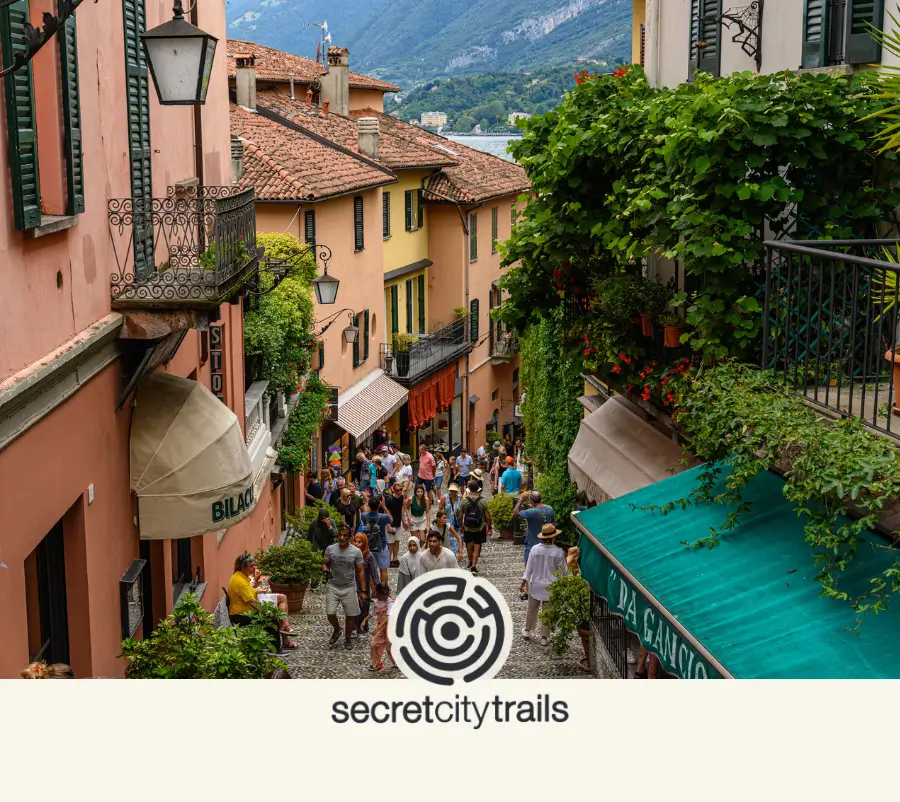 Crowds walking up Salita Serbelloni, a picturesque staircase lined with colorful buildings in Bellagio with the secretcitytrails logo